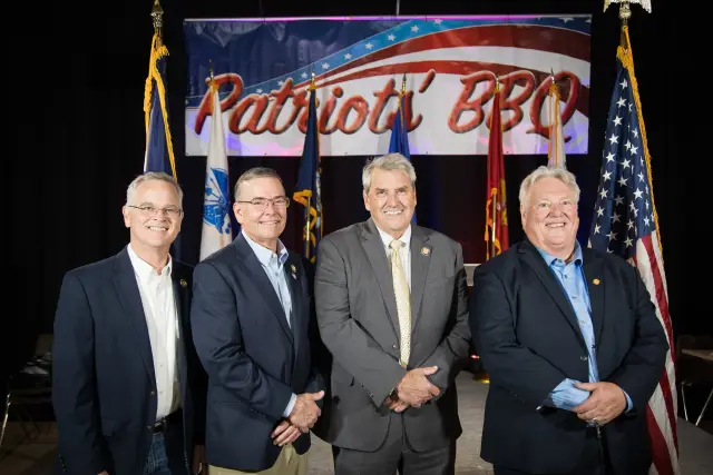 Four men pose under the Patriots' BBQ banner